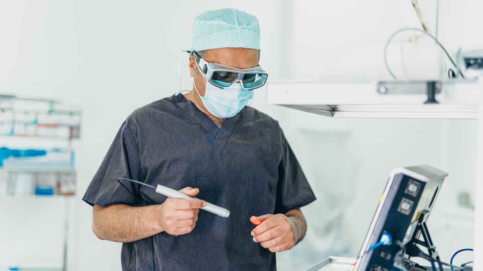 Herr Doctor-medic Naes trägt einen Kasack, eine Schutzmaske, Schutzbrille und OP-Haube. Mit der rechten Hand prüft er das rote Licht eines Lasers an der anderen Hand. Rechts am Rand ist noch seitlich der Monitor des Behandlungsgeräts zu sehen.