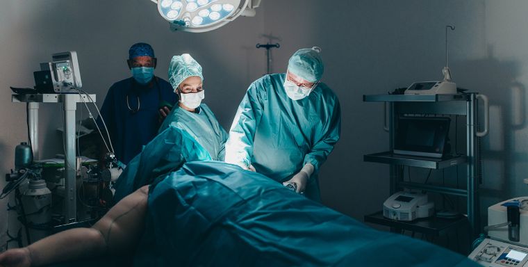 Aufnahme einer OP-Situation. Der Raum ist dunkel, nur der Behandlungsbereich ist erleuchtet. Drei Personen stehen in voller OP-Montur um einen Patienten, der auf dem OP-Tisch liegt. Links und rechts sind medizinische Geräte zu sehen.