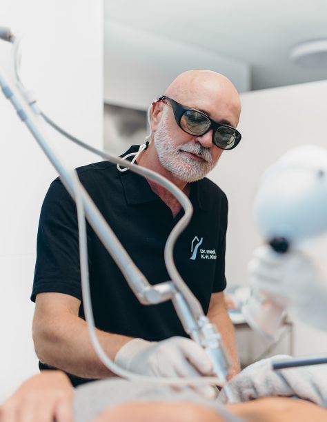 Herr Dr. Klein ist in eine Behandlung vertieft. Er trägt eine Schutzbrille und Handschuhe und behandelt eine nicht sichtbare Person mit einem Lasergerät. Dr. Klein trägt das T-Shirt der Praxis.