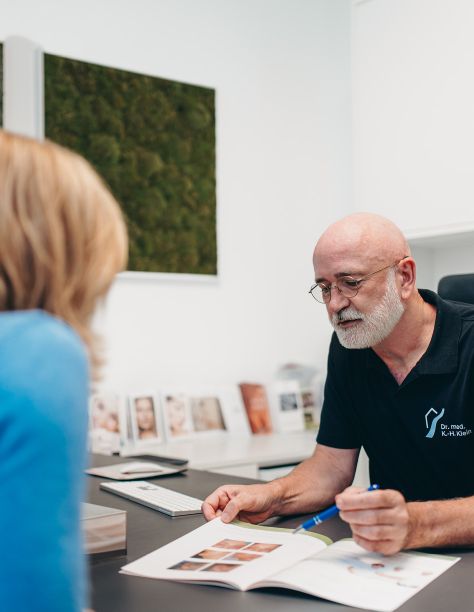 Foto von Dr. Klein, der am Schreibtisch sitzt und in einer Broschüre blättert. Im Gegenüber sitzt eine Patientin mit blauen Oberteil. Der Hintergrund ist verschwommen.