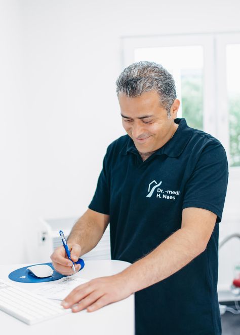 Herr Doctor-medic Naes steht an einem Stehtisch und schreibt auf einem Zettel. Er trägt das dunkelblaue Praxis-Shirt der Praxisklinik. Ein Behandlungszimmer ist verschwommen im Hintergrund zu sehen.