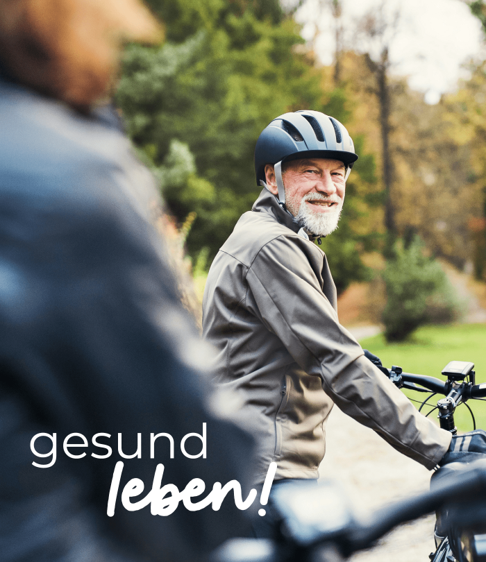 Ein älterer Herr mit weißem Bart sitzt auf einem Fahrrad und blickt zu der Person im Vordergrund, die verschwommen zu sehen ist. Er lächelt und trägt einen Helm und eine Outdoor-Jacke. Auf dem Bild steht "gesund leben!".