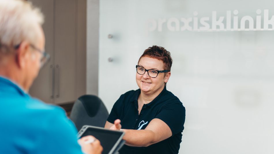 Eine Sprechstundenhilfe am Empfang reicht einem männlichem Patienten ein Tablet zum Ausfüllen von Daten. Sie hat kurzes rötliches Haar, trägt eine Brille und ein dunkelblaues T-Shirt. Im Hintergrund ist das Wort "Praxisklinik" zu lesen.