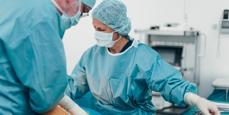 Zwei Personen in hellblauer OP-Kleidung, mit Hauben, Handschuhen und Schutzmasken sind in eine Behandlungssituation vertieft. Im Hintergrund sind verschwommen Behandlungsgeräte zu sehen.