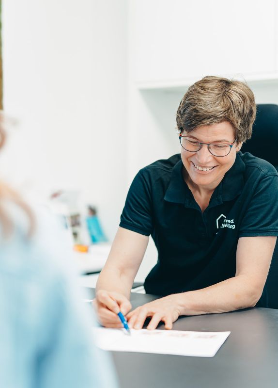 Frau Dr. Nölling sitzt lächelnd am Schreibtisch und zeigt einer verschwommenen Person im Vordergrund mit dem Kugelschreiber etwas in einer Broschüre, die vor ihr liegt.