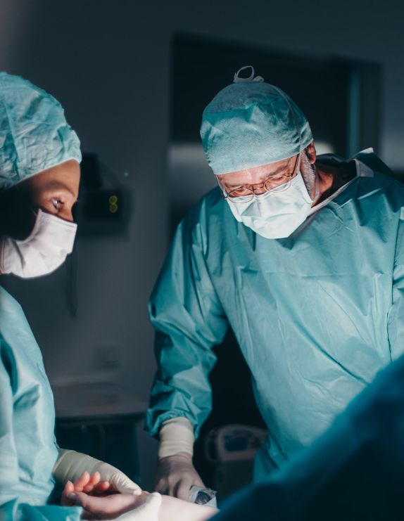 Ein dunkler Raum, nur die OP-Situation ist hell erleuchtet. Dr. Klein (rechts) und eine Frau (links) tragen hellblaue OP-Kleidung mit Masken, Hauben und Handschuhen und beugen sich zu einem Patienten runter.