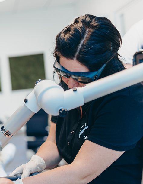 Eine schwarzhaarige MFA ist in eine Behandlung vertieft. Sie trägt eine Schutzbrille und Handschuhe, ein Metallteil eines Behandlungsgeräts verläuft durch das Bild. Der Hintergrund ist verschwommen.