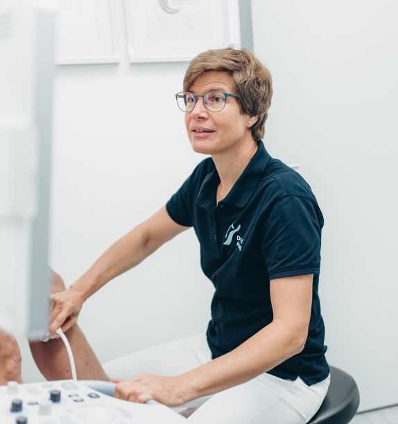 Frau Dr. Nölling,blickt auf einen Monitor, der links am Bildrand zu sehen ist. Mit der rechten Hand untersucht sie mit einem gerät das Bein eines Patienten. Sie trägt ein dunkelblaues Praxis-Shirt.