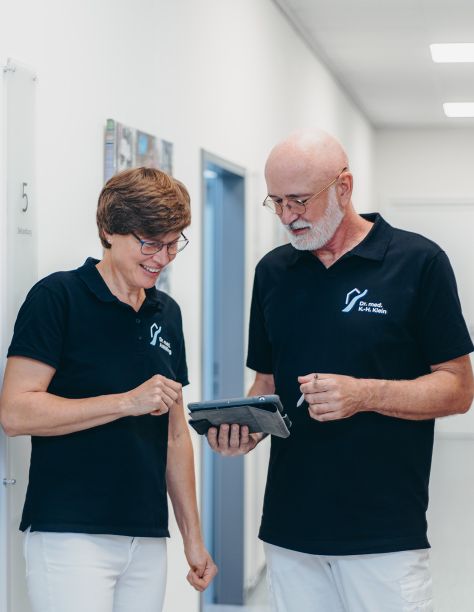 Frau Dr. Nölling und Herr Dr. Klein, stehen nebeneinander im Flur der Praxis und blicken gemeinsam auf ein Tablet.
