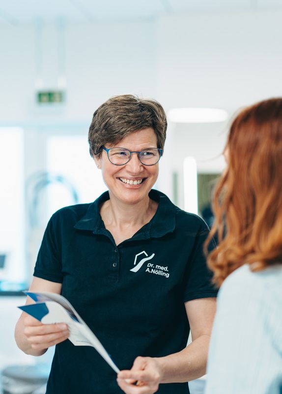 Frau Dr. Nölling lächelt eine rothaarige Frau, vermutlich eine Patientin, an. In der Hand hält sie eine Broschüre der Praxis, sie trägt das dunkelblaue Praxis-Shirt der Praxisklinik am Flüsschen. Der Hintergrund ist verschwommen.