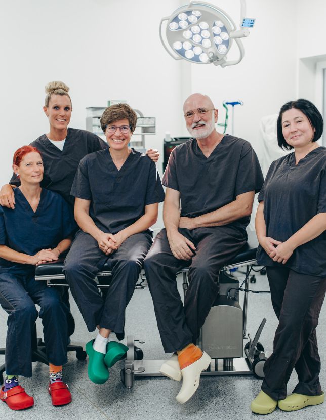 Ein Gruppenbild mit Dr. Klein und Dr. Nölling, die auf einer Behandlungsliege sitzen. Links neben Dr. Nölling sind zwei MFA, eine sitzt und die andere steht hinter den beiden. Rechts von Dr Klein steht eine MFA. Alle tragen dunkle OP-Kleidung.
