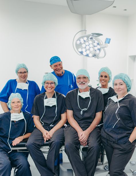Gruppenbild des OP-Teams. Alle tragen dunkle Kasacks, Op-Hauben und die Masken liegen offen am Hals der 7 Personen. Dr. Nölling und Dr. Klein sitzen in der Mitte auf einer Liege.
