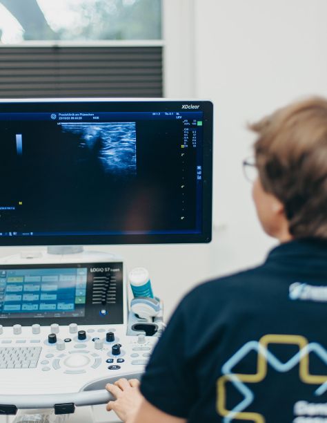 Frau Dr. Nölling von hinten, verschwommen im Vordergrund, blickt im Hintergrund auf einen dunklen Monitor, auf dem eine Ultraschallaufnahme zu sehen ist.