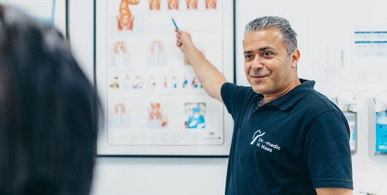 Herr Doctor-medic Naes steht vor einem Schaubild mit schematischen Darstellungen der Enddarmregion und zeigt einer nicht sichtbaren Person die Bilder. Er trägt das dunkelblaue Praxis-Shirt der Praxisklinik.
