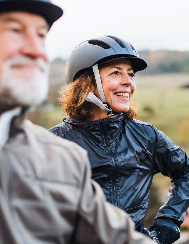 Ein älteres Paar ist auf Fahrradtour unterwegs. Der Mann, verschwommen im Vordergrund, hat einen grauen Bart, trägt einen Helm und eine dunkle Jacke. Die Frau im Hintergrund lächelt, trägt einen Helm und eine schwarze Jacke.