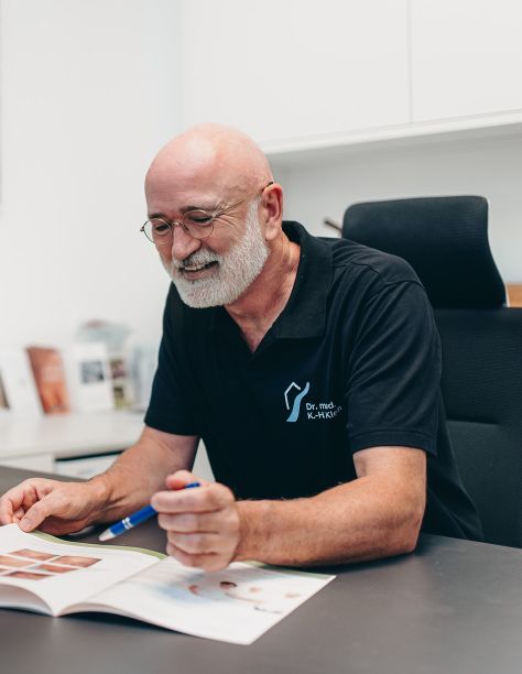Foto von Dr. Klein, der am Schreibtisch sitzt und in einer Praxis-Broschüre blättert. Er lächelt herzlich. Sein Oberkörper wird von dem Stilelement der Praxisklinik am Flüsschen umrahmt.