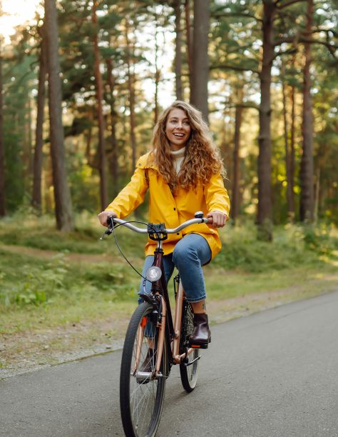 Eine junge Frau mit blonden, langen Haaren und gelber Jacke, fährt auf einem Fahrrad im Wald.