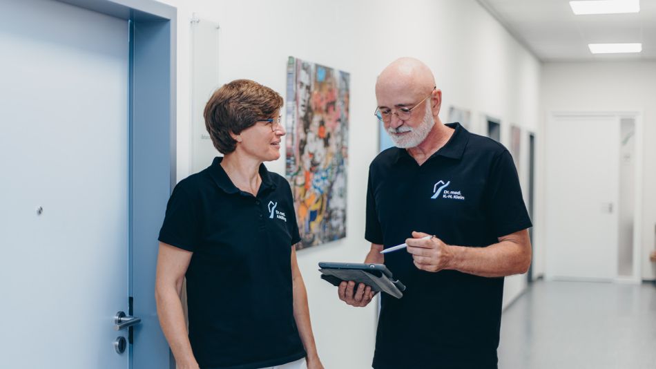 Frau Dr. Nölling und Herr Dr. Klein, stehen nebeneinander im Flur der Praxis. Dr. Klein blickt auf ein Tablet. Dr. Nölling sieht Dr. Klein an. Sie tragen dunkelblaue T-Shirts.