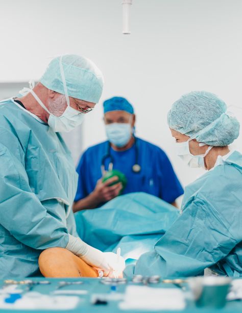 Herr Dr. Klein und eine weitere Person befinden sich im OP. Alle tragen OP-Montur, mit Hauben, Schutzmasken und Handschuhen. Eine Person wird am Bein zur Behandlung eines Lipödems operiert.