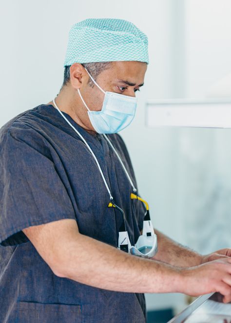 Herr Doctor-medic Naes trägt einen Kasack, eine Schutzmaske und OP-Haube. Die Schutzbrille liegt mit einem Band verbunden auf seiner Brust.Er blickt konzentriert auf einen Monitor.