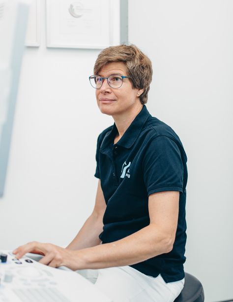 Frau Dr. Nölling sitzt entspannt auf einem Hocker und bedient ein Behandlungsgerät. Sie blickt vermutlich auf den Monitor, der auf dem Bild nicht mehr zu sehen ist. Sie trägt ein dunkelblaues T-Shirt und eine weiße Hose.