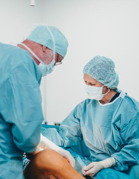 Zwei Personen in hellblauer OP-Kleidung, mit Hauben, Handschuhen und Schutzmasken sind in eine Behandlungssituation vertieft. Im Hintergrund sind verschwommen Behandlungsgeräte zu sehen.
