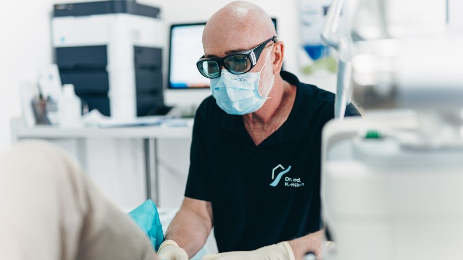 Aufnahme von Herrn Dr. Klein. Er trägt das dunkelblaue Praxis-Shirt, eine Schutzbrille, eine Schutzmaske und Handschuhe. Der Hintergrund ist verschwommen und zeigt grob eine Behandlungssituation in der Praxis.