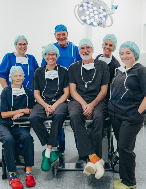 Teamfoto des OP-Teams. Es sind 7 lachende Personen im Kasack und OP-Haube zu sehen, das Foto wurde auch in einem OP-Raum aufgenommen. Frau Dr. Nölling und Herr Dr. Klein sitzen dabei in der Mitte auf einerOP-Liege, umrandet von Ihren Kollegen, die stehen.