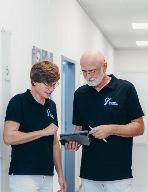 Frau Dr. Nölling und Herr Dr. Klein, stehen nebeneinander im Flur der Praxis und blicken gemeinsam auf ein Tablet.