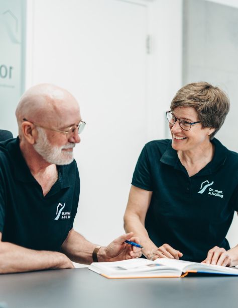 Herr Dr. Klein (rechts) und Frau Dr. Nölling (links) sitzen an einem Tisch und lachen sich an. Vor ihnen liegt ein aufgeschlagenes Buch. Beide tragen die Praxis-T-Shirts.