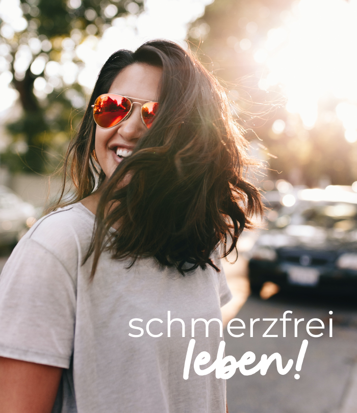 Eine junge Frau lacht in die Kamera. Ihr braunes, schulterlanges Haar wird in ihr Gesicht geweht. Sie lacht und trägt eine rote Sonnenbrille und ein graues T-Shirt. Auf dem Bild steht "schmerzfrei leben!".