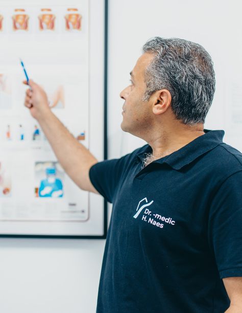Herr Doctor-medic Naes blickt auf ein Schaubild an der Wand und deutet mit der rechten Hand darauf. Es sind schematische Darstellungen des Körpers zu sehen. Er trägt das dunkelblaue Praxis-Shirt der Praxisklinik.