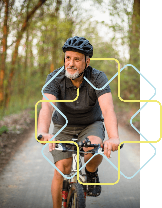 Ein älterer Mann mit grauem Bart fährt auf einem Mountainbike auf einem Weg im Wald. Er trägt einen Helm, ein dunkelgraues T-Shirt und eine dunkle kurze Hose. Das geometrische Stilelement in gelb und hellblau der Praxisklinik umrahmt seinen Oberkörper.