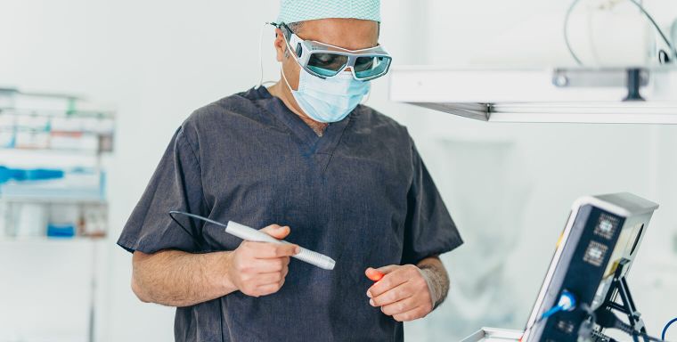 Herr Doctor-medic Naes trägt einen Kasack, eine Schutzmaske, Schutzbrille und OP-Haube. Mit der rechten Hand prüft er das rote Licht eines Lasers an der anderen Hand. Rechts am Rand ist noch seitlich der Monitor des Behandlungsgeräts zu sehen.