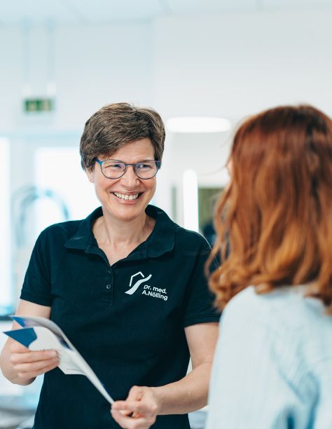 Frau Dr. Nölling lacht eine Person links von ihr an. Sie steht rechts und trägt das T-Shirt der Praxis. In der Hand hält sie eine Informationsbroschüre der Praxis. Der Hintergrund ist verschwommen.