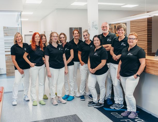Ein Gruppenbild von 10 Mitarbeitern der Praxis im Empfangsbereich der Praxisklinik im Burbach. Alle Personen tragen weiße Hosen und die Praxis-T-Shirts. Unter den Personen sind Dr. Klein und Dr. Nölling.